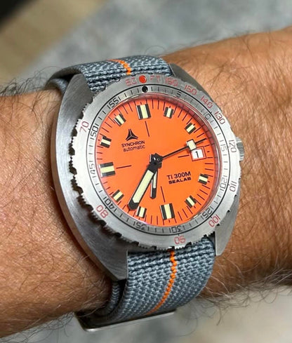 Bracelet parachute élastique style PLD Marine française gris anthracite avec bande orange, installé sur une montre de plongée vintage au poignet.